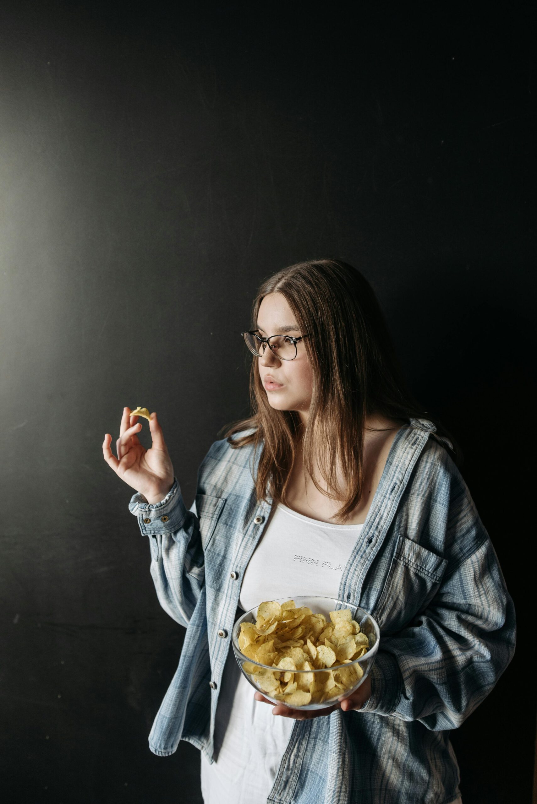 Young woman with glasses holding a bowl of chips in a relaxed indoor setting.